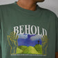 Jade Green Garment Dyed T-Shirt with White Text(BEHOLD) over green pasture with hands on sides.