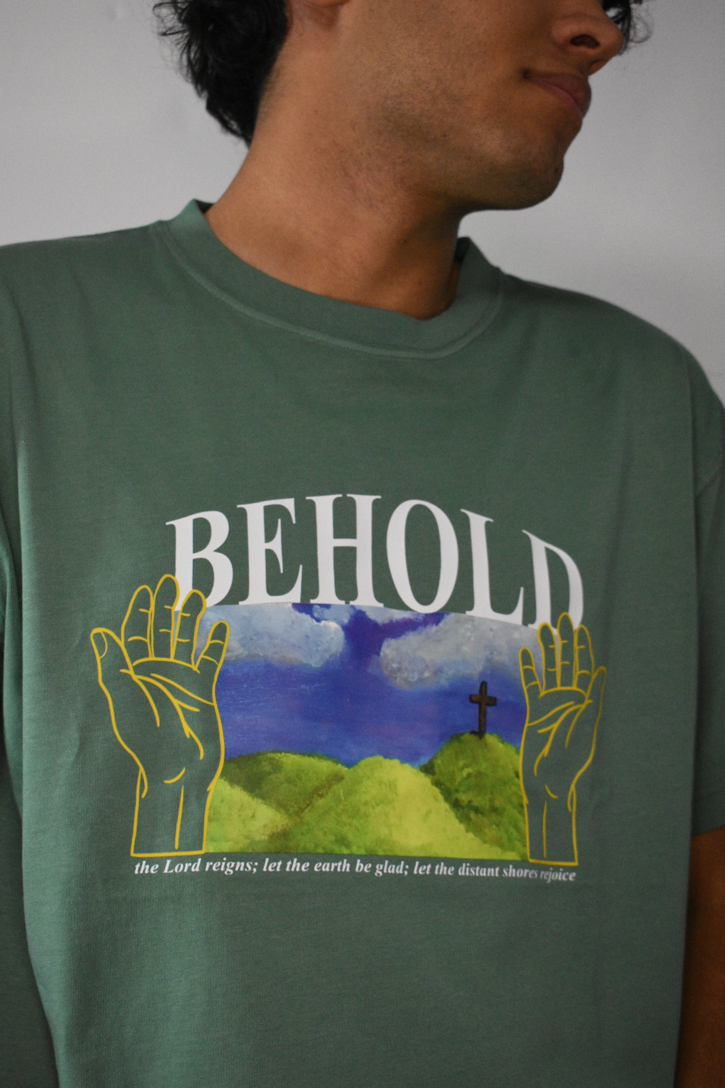 Jade Green Garment Dyed T-Shirt with White Text(BEHOLD) over green pasture with hands on sides.